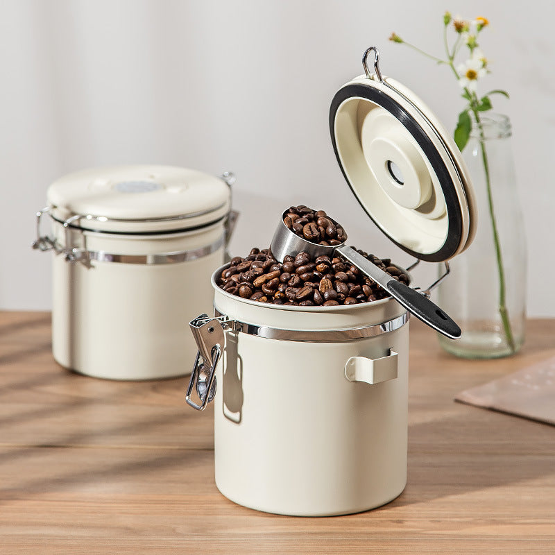 White coffee canister with open lid, beans, and scoop on a wooden surface.